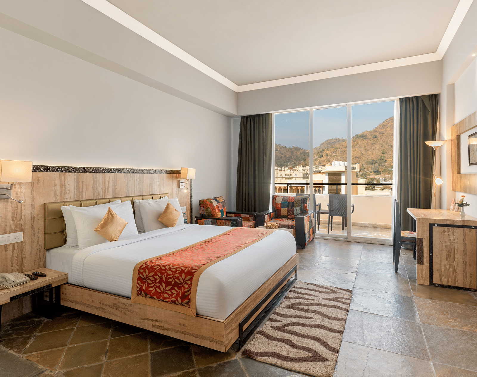 A hotel room with a bed, seating, desk, a large window leading to a balcony, and a patterned rug - Via Lakhela Resort & Spa, Kumbhalgarh