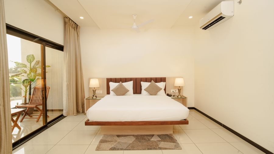 Hotel room with a clean wooden bed frame and white bedding, featuring bright daylight entering through a large sliding door at Arawali Ananta Elite, Jaipur.