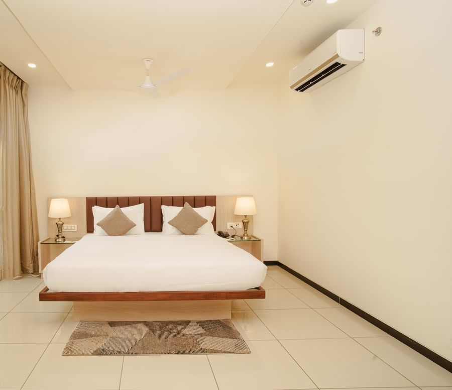 Minimalist hotel room with a wooden frame bed, white walls, and a glass door opening to a bright outdoor view at Arawali Ananta Elite, Jaipur.