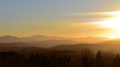 A golden sunset casting warm light over layered hills and distant mountains, creating a serene, scenic view. 