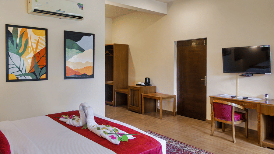 Spacious guest room at Infinity Resorts Corbett featuring a double bed with decorative towel art, a wall mounted TV, a work desk, air conditioning, and contemporary artwork.