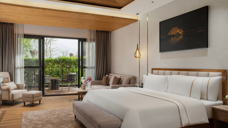 A bright hotel room with a sofa set, double bed, and large windows revealing a beautiful mountain landscape.