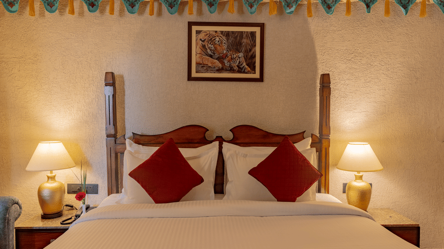 Close-up of a bed with pillows and bedside lamps at Estherea Bagh, Ranthambore, featuring a framed picture and colourful wall hangings above.