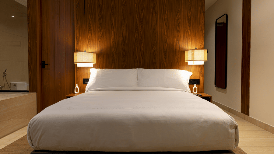 Hotel room at SUKAVI with a large bed dressed in white linens, a dark wooden headboard, and 2 bedside lamps turned on.