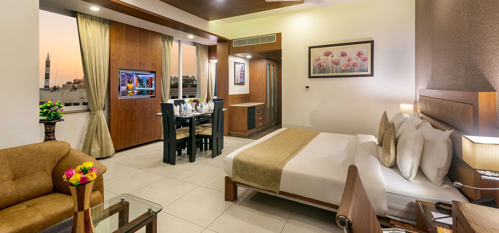 A spacious hotel room with a bed, seating area with a couch and glass table, dining table, and a view outside the window.