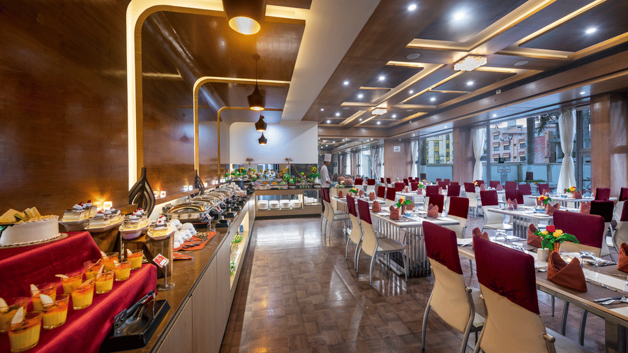 A restaurant with modern decor, food counters, and neatly set dining tables featuring a long counter displaying assorted desserts and dishes at Lords Inn Somnath