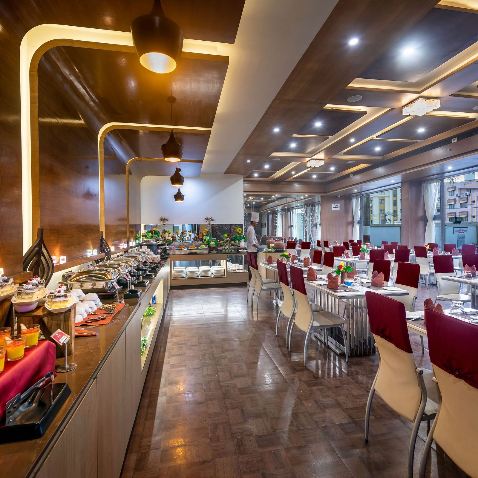 A restaurant with modern decor, food counters, and neatly set dining tables featuring a long counter displaying assorted desserts and dishes at Lords Inn Somnath