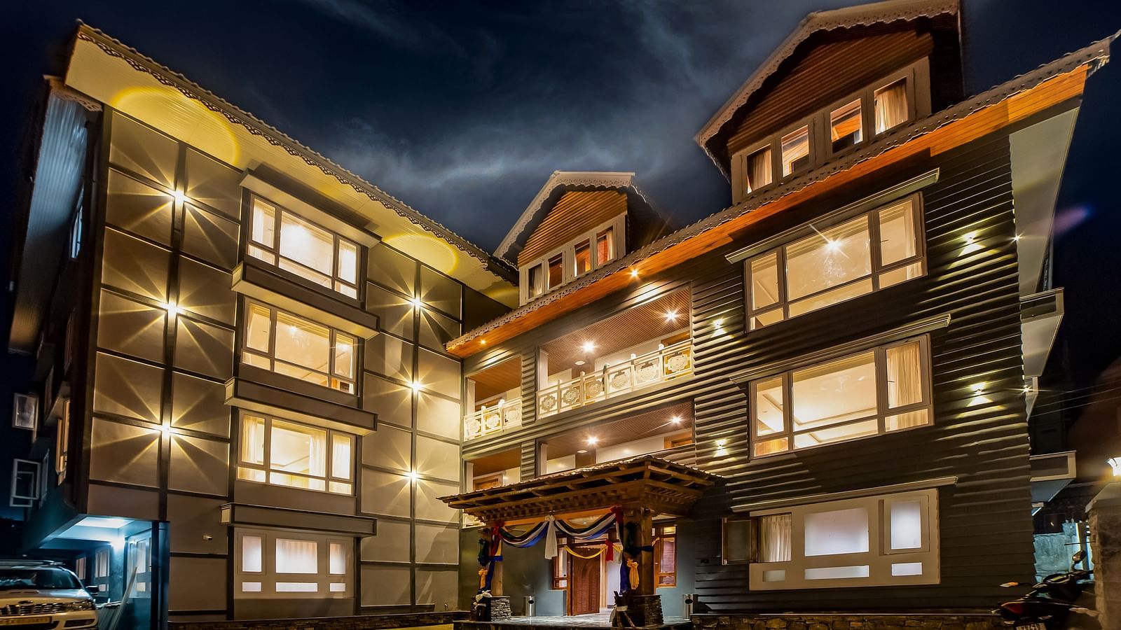 Well-lit modern hotel exterior at night with large illuminated windows, a central balcony, decorative entrance with drapes, and a paved driveway under a dramatic evening sky - Yashshree Lachen.