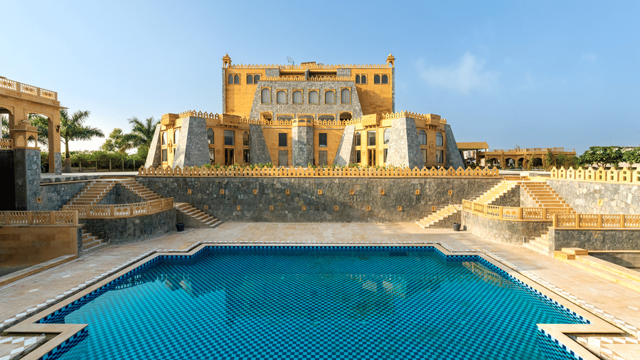 An outdoor swimming pool at EsthereaRaj Leela, Ranakpur with a stone building facade in the background under a blue sky.