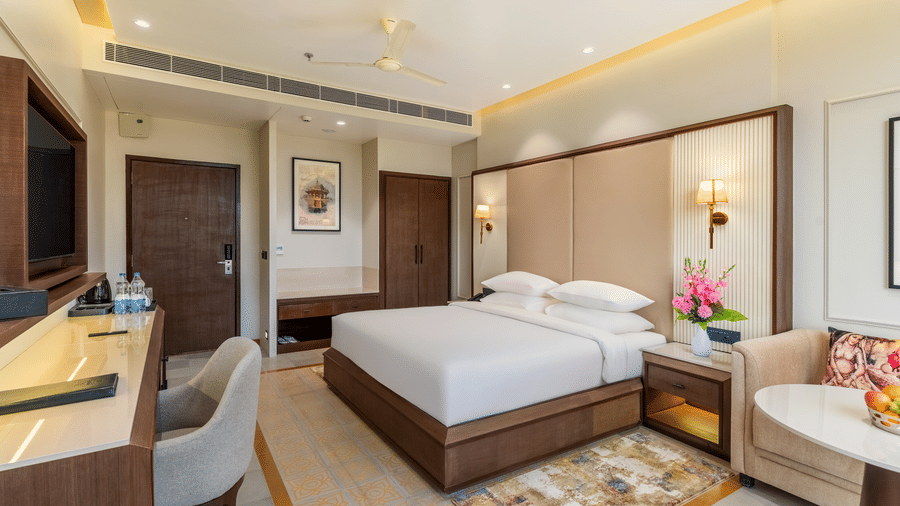 A wide view of a room at EsthereaRaj Leela, Ranakpur, featuring a king size bed with a tan headboard, a work desk, a wall-mounted TV, and a side seating area.