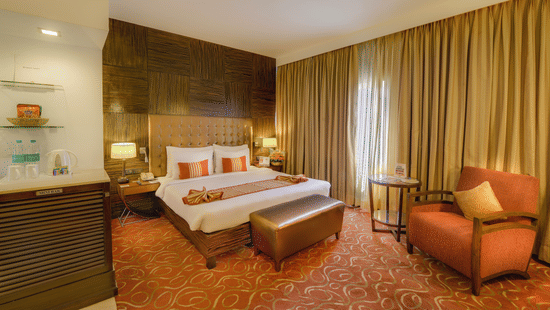 Premium Room at Pride Premier Chennai featuring a plush bed with a diwan at the foot and a couch placed next to the large window.