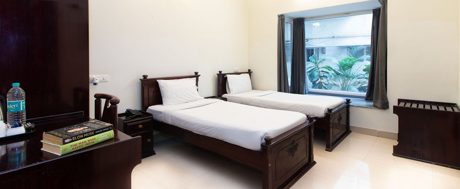 A room offering twin beds with white linens, wooden furniture including a wardrobe, a table, a chair, and a window letting in natural light at Sanctum Suites Indiranagar, Bangalore.