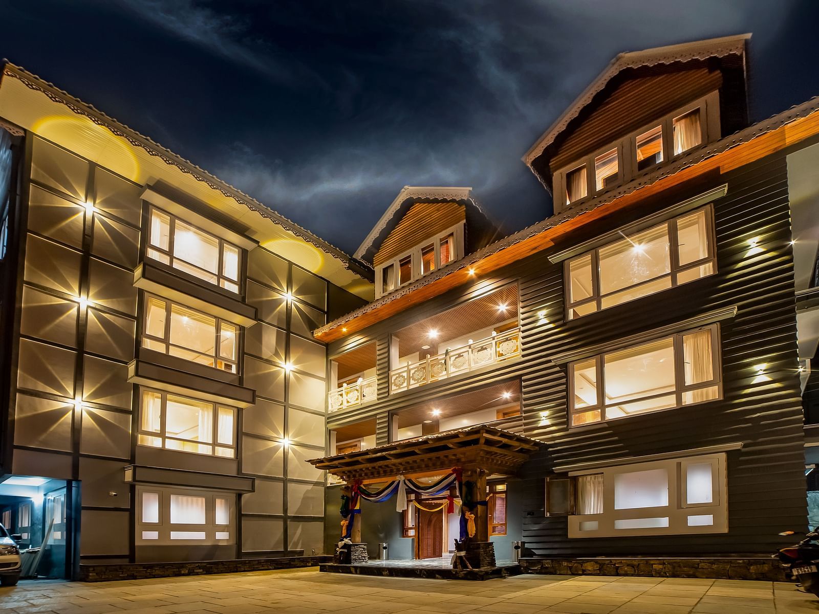 Well-lit modern hotel exterior at night with large illuminated windows, a central balcony, decorative entrance with drapes, and a paved driveway under a dramatic evening sky - Yashshree Lachen.