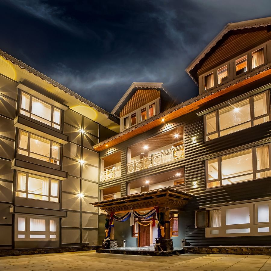 Modern hotel building at night with warm lighting, large windows, a central balcony, and a decorative entrance with drapes, set under a dramatic, cloudy evening sky – Yashshree Lachen.
