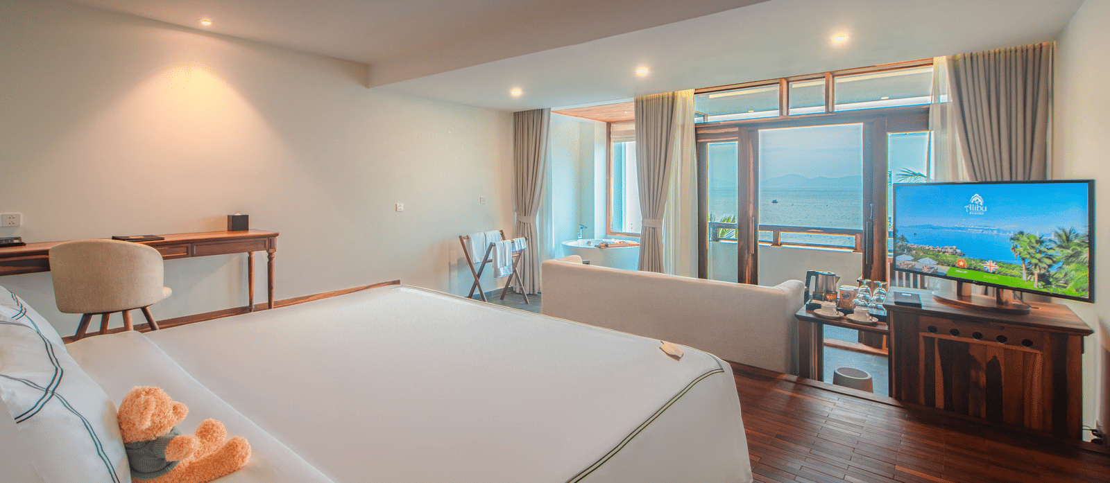 Executive Ocean Suite at Alibu Resort Nha Trang, Vietnam, featuring king bed with white linens, seating area, writing desk, wide windows, wooden flooring and TV near patio doors.