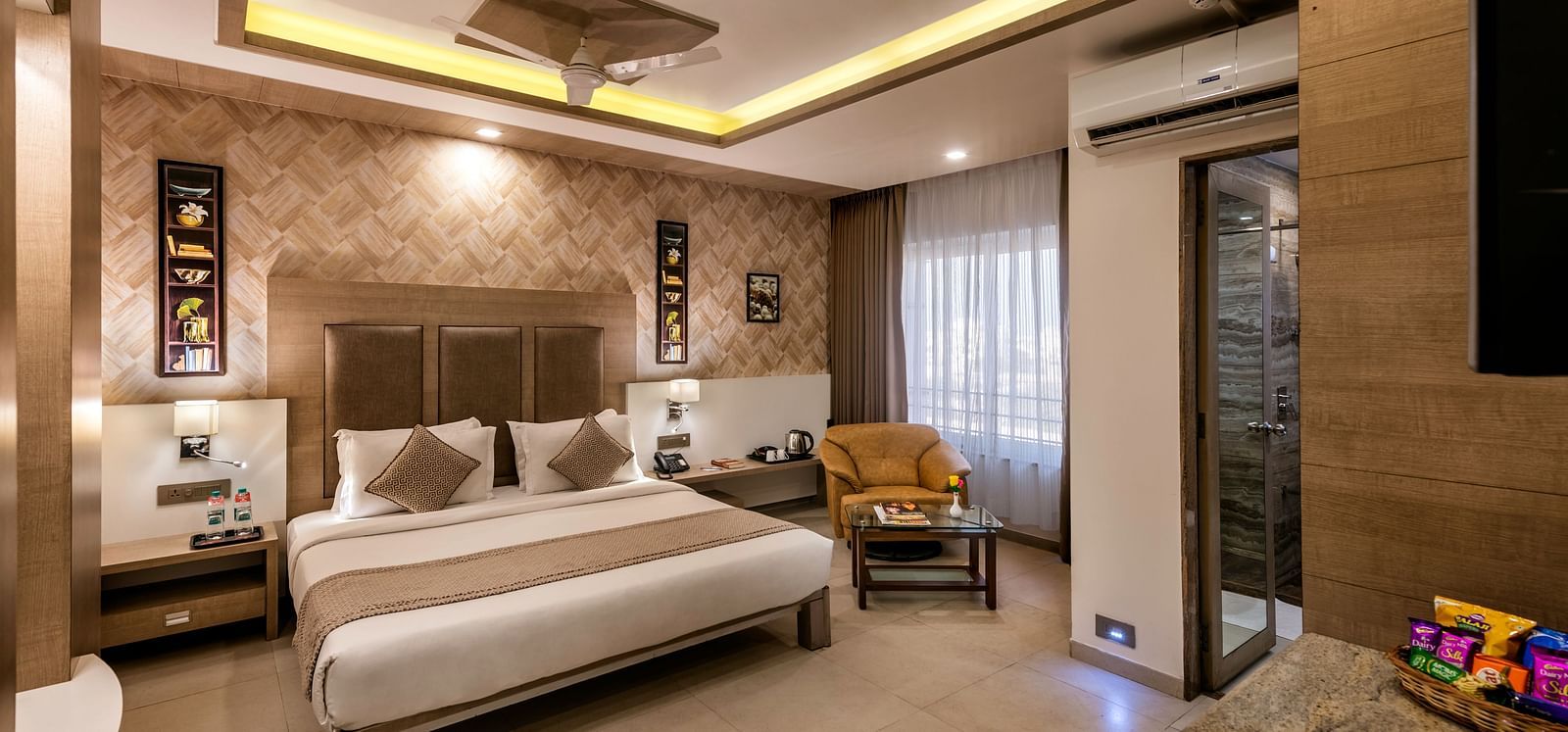 room with a king-sized bed adorned with neutral-toned linens and pillows, a comfortable armchair and small coffee table situated near a window with sheer curtains, ambient lighting highlighting patterned wallpaper, and a glimpse into an en-suite bathroom a