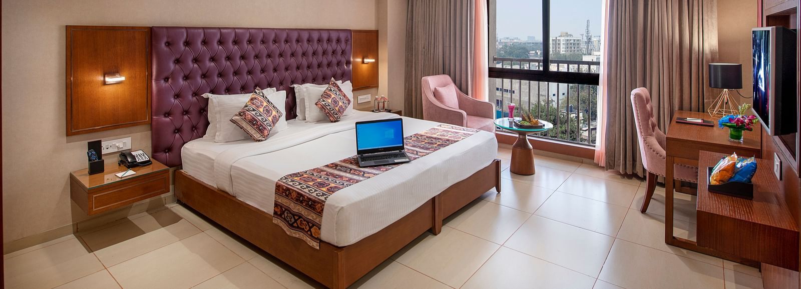 A bright hotel room featuring a large bed with a purple headboard, a work desk and city views from the window -  SK Lords Inn Ahmedabad