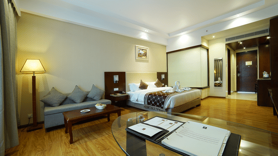 Elegant hotel suite featuring a double bed, a grey sofa with cushions, a wooden coffee table, and warm lighting on polished wooden floors at Golden Tulip Kukas, Jaipur.