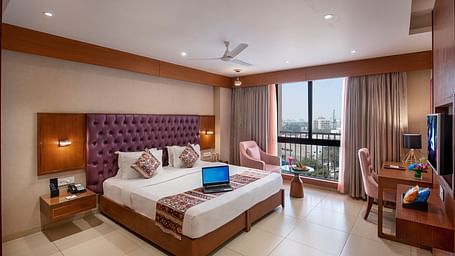 A bright hotel room featuring a large bed with a purple headboard, a work desk and city views from the window -  SK Lords Inn Ahmedabad