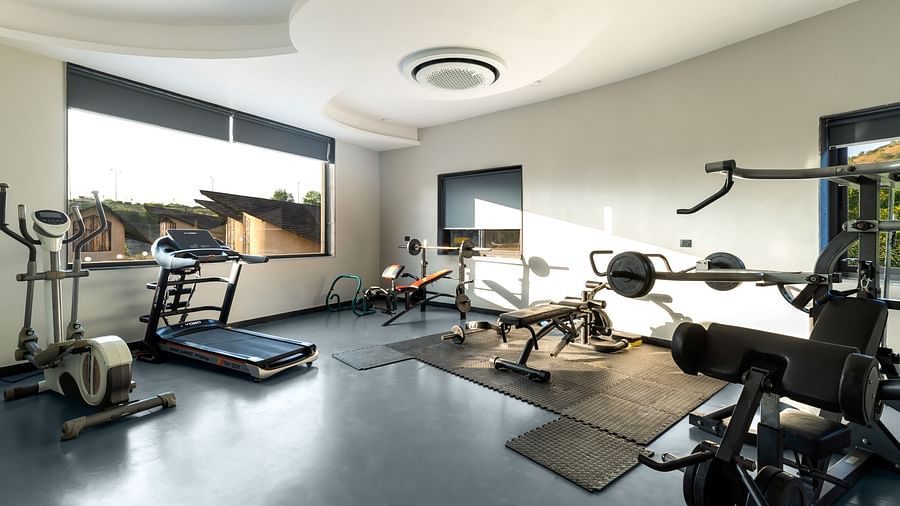 A well-equipped gym at The Qualia Resort Club & Brewery, Udaipur, complete with treadmills, weight machines, and large windows offering natural light.