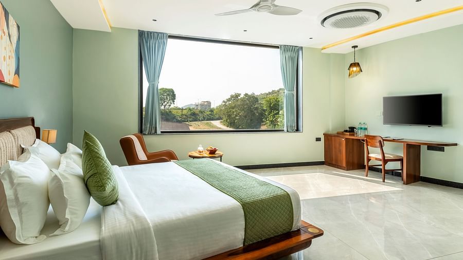 Pool View Room presenting a king size bed with white and green linens, a ceiling fan, and a view of greenery through a large window at The Qualia Resort Club & Brewery, Udaipur.