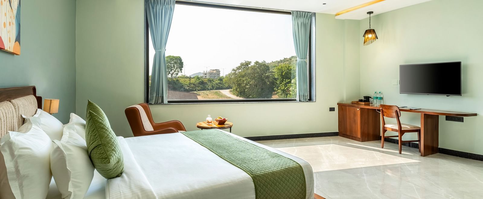 Pool View Room presenting a king size bed with white and green linens, a ceiling fan, and a view of greenery through a large window at The Qualia Resort Club & Brewery, Udaipur.