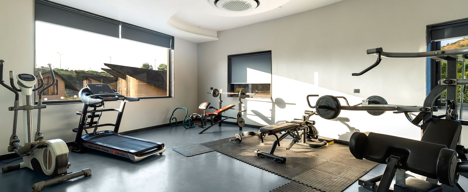 A well-equipped gym at The Qualia Resort Club & Brewery, Udaipur, complete with treadmills, weight machines, and large windows offering natural light.