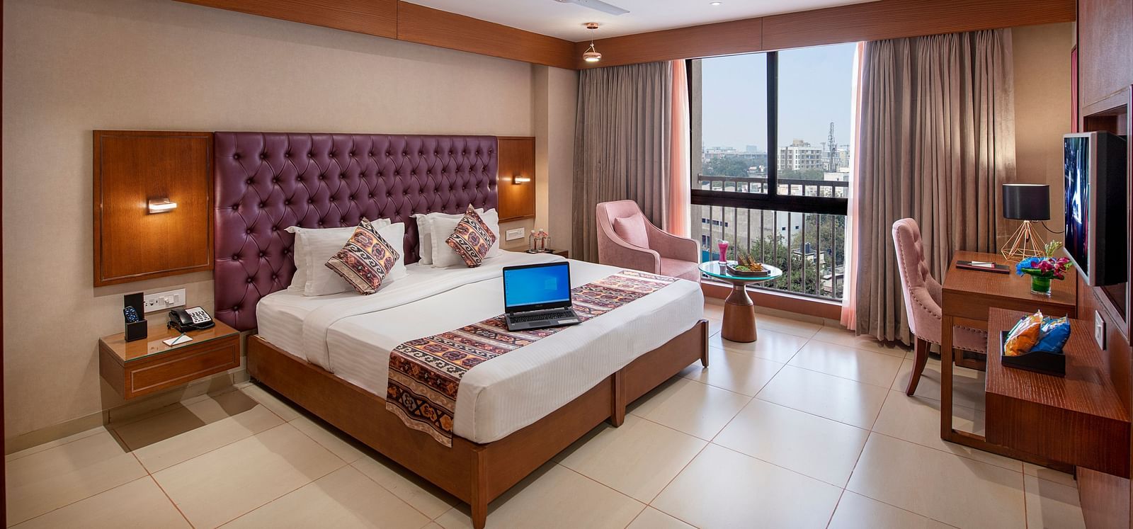 A bright hotel room featuring a large bed with a purple headboard, a work desk and city views from the window -  SK Lords Inn Ahmedabad