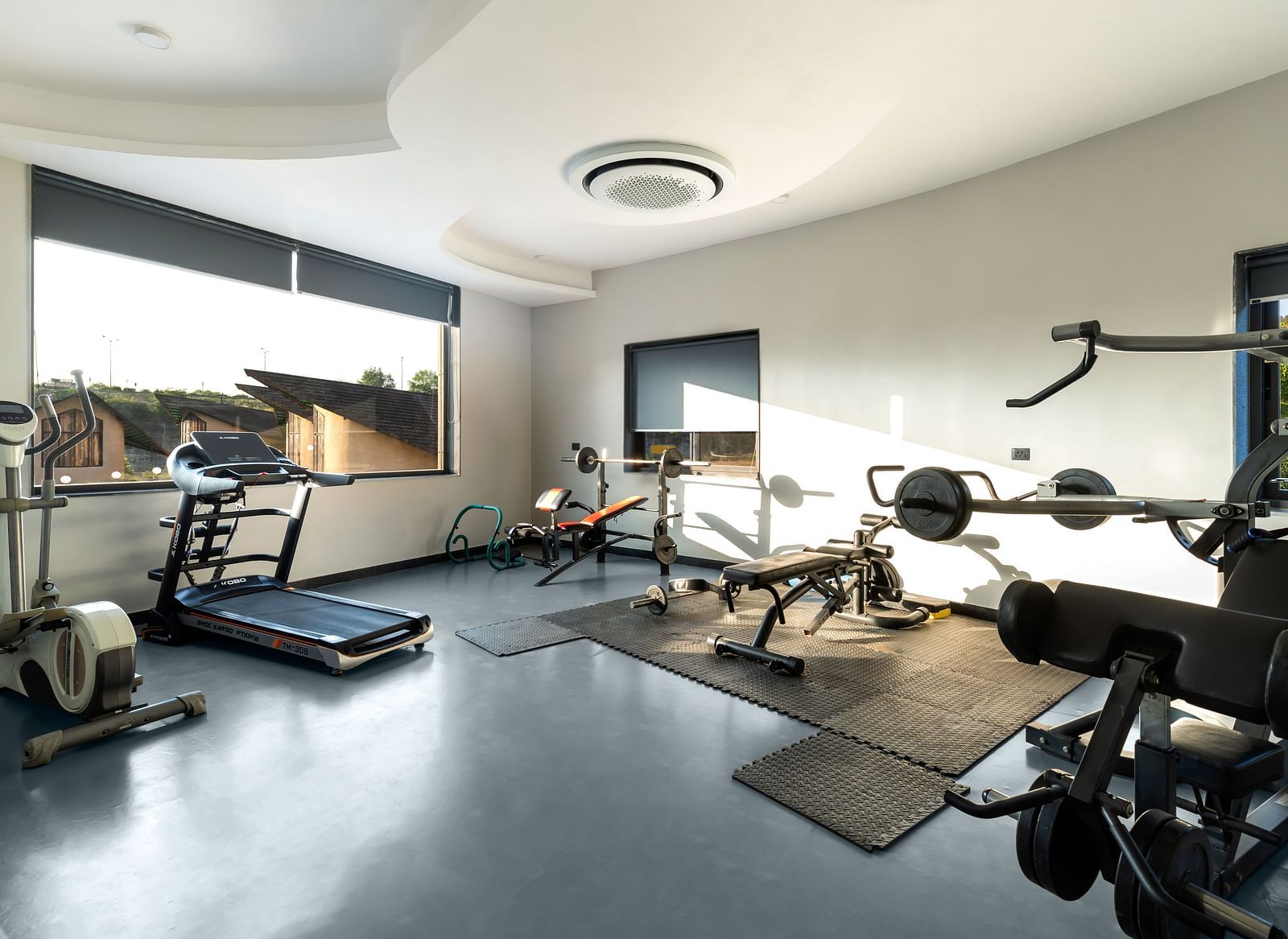 A well-equipped gym at The Qualia Resort Club & Brewery, Udaipur, complete with treadmills, weight machines, and large windows offering natural light.