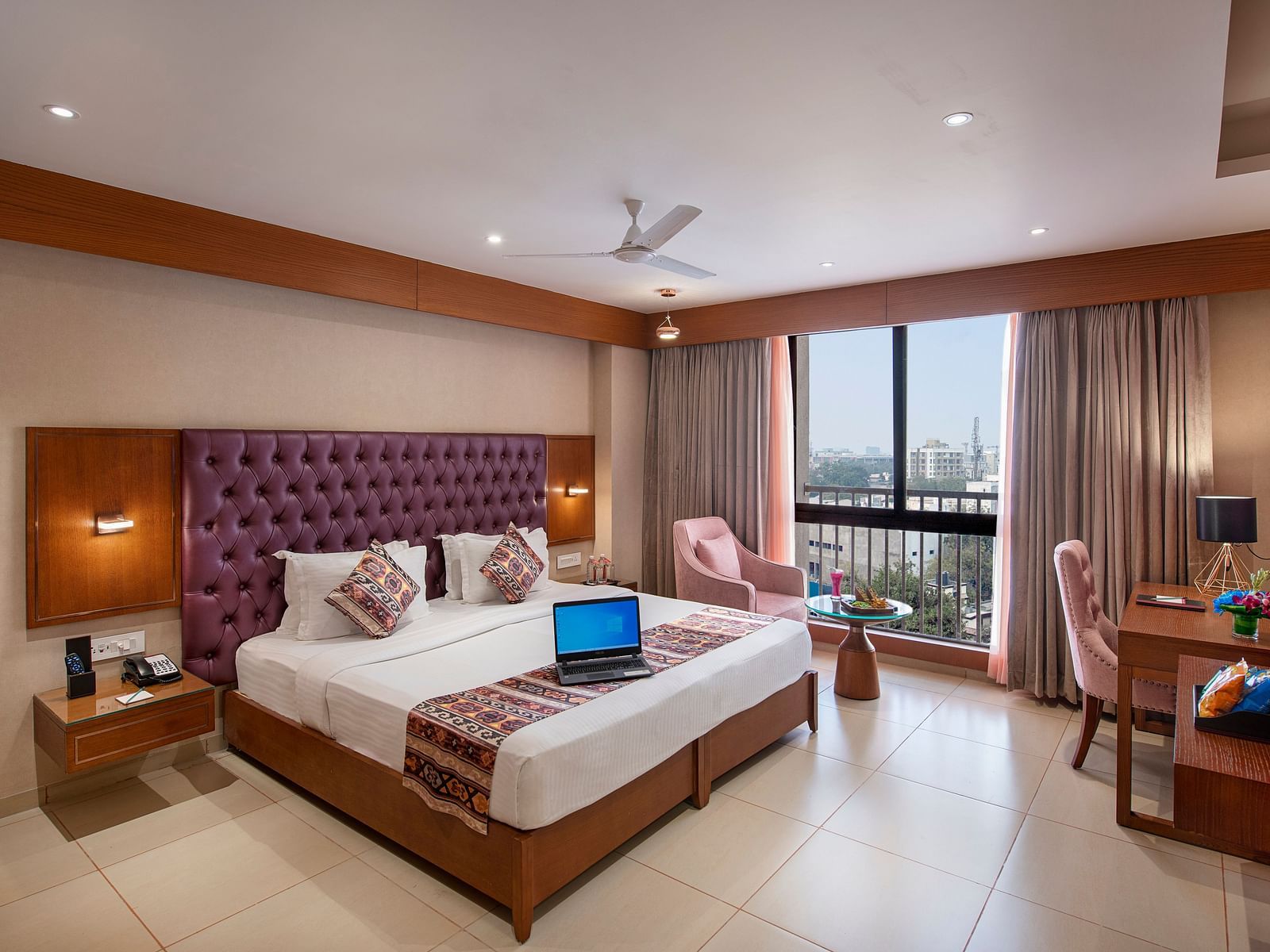 A bright hotel room featuring a large bed with a purple headboard, a work desk and city views from the window -  SK Lords Inn Ahmedabad