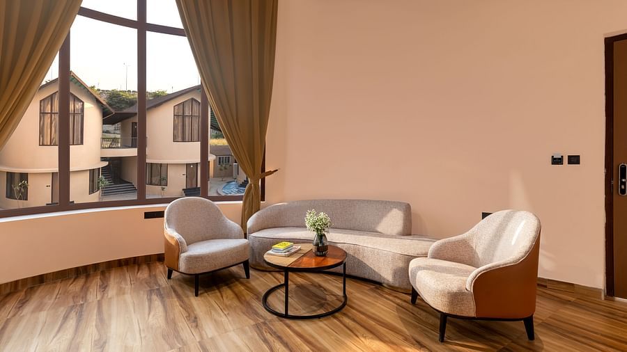 Luxury Villa with Sitout features a seating area with a curved sofa, two armchairs, and a view of the outdoors through large windows at The Qualia Resort Club & Brewery, Udaipur.