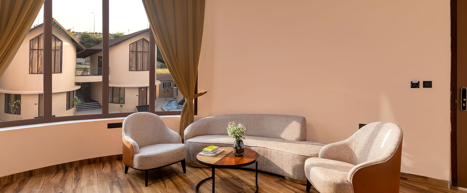 Luxury Villa with Sitout features a seating area with a curved sofa, two armchairs, and a view of the outdoors through large windows at The Qualia Resort Club & Brewery, Udaipur.