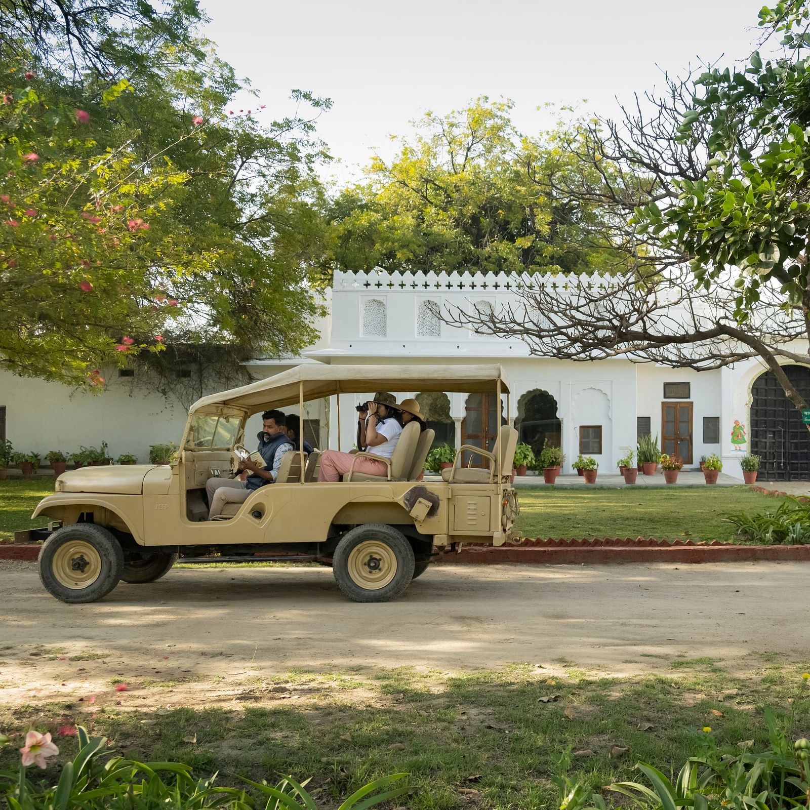 A beige utility vehicle with passengers parked in front of a white building with a flat roof, surrounded by trees and greenery- Shahpura Bagh.