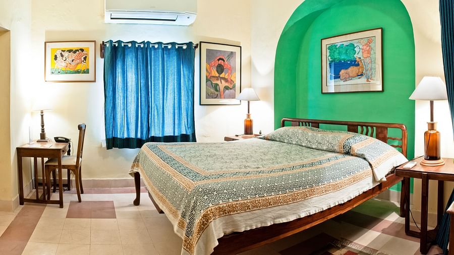 Bedroom with a bed featuring a patterned cover, bedside tables with lamps, a desk and chair, artwork, blue curtains, and AC at Neemrana's Tijara Fort Palace, Alwar.