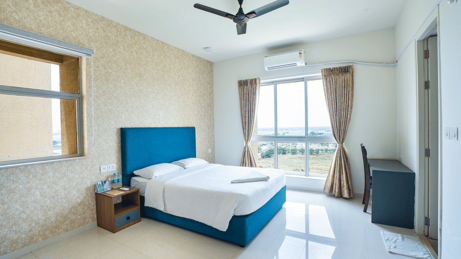 A minimalist bedroom with a blue upholstered bed, a textured gold feature wall, and a large window with neutral-toned curtains at Apartel by Aarin, Oragadam.