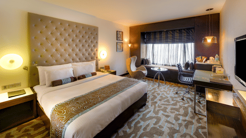Superior Twin Room at Pride Premier Bengaluru featuring a plush bed with a cushiony headboard in front of a TV mounted on the wall and a sitting space is seen next to the window.