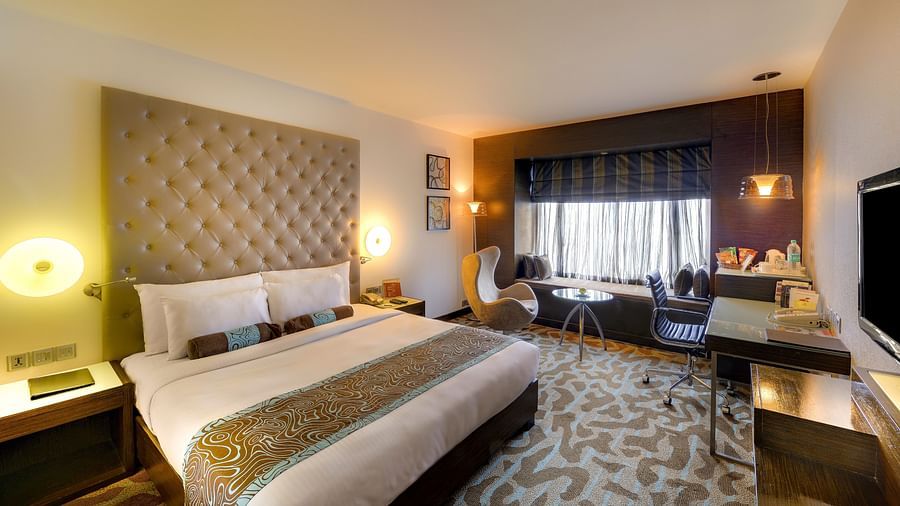 Superior Room at Pride Hotel, Bangalore featuring a plush bed with a cushiony headboard in front of a TV mounted on the wall and a sitting space is seen next to the window
