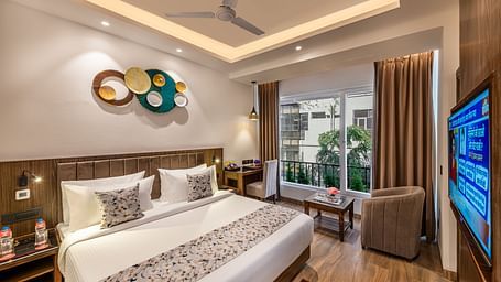 A bright, modern hotel room with a king size bed, decorative wall art, a brown armchair, small table, flat-screen TV, and an open door leading to a balcony - Lords Eco Inn Dehradun.