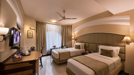 Modern designed minimalistic room with a twin bed, a plush armchair and a table, bedside lamps, mounted T.V., and floor-to-ceiling window with curtains at Lords Inn Vadodara.