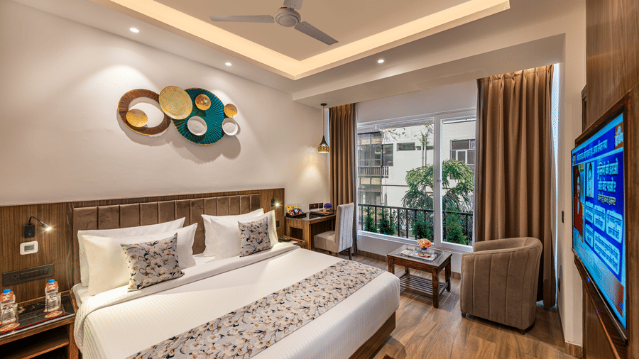 A bright, modern designed room with a king size bed, bedside table, decorative wall art, a brown armchair, small table, flat-screen TV, and a glass door leading to a balcony at Lords Eco Inn Dehradun.