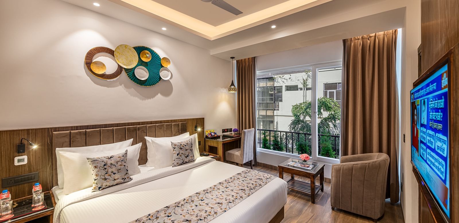 A bright, modern hotel room with a king size bed, decorative wall art, a brown armchair, small table, flat-screen TV, and an open door leading to a balcony - Lords Eco Inn Dehradun.
