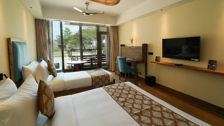 Modern hotel room with two twin beds, a wooden desk with a blue chair, a wall-mounted TV, and a private balcony overlooking a green outdoor space - MGM Beach Resorts