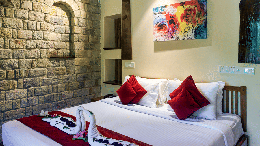 Comfortable guest room at Infinity Resorts Corbett featuring a double bed with decorative towel art, a stone accent wall, bedside lighting, and contemporary artwork.