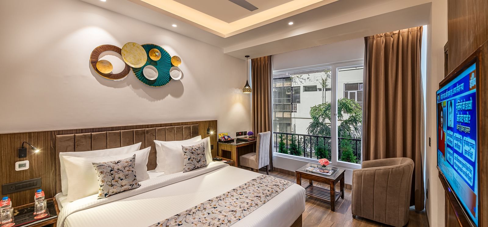 A bright, modern hotel room with a king size bed, decorative wall art, a brown armchair, small table, flat-screen TV, and an open door leading to a balcony - Lords Eco Inn Dehradun.
