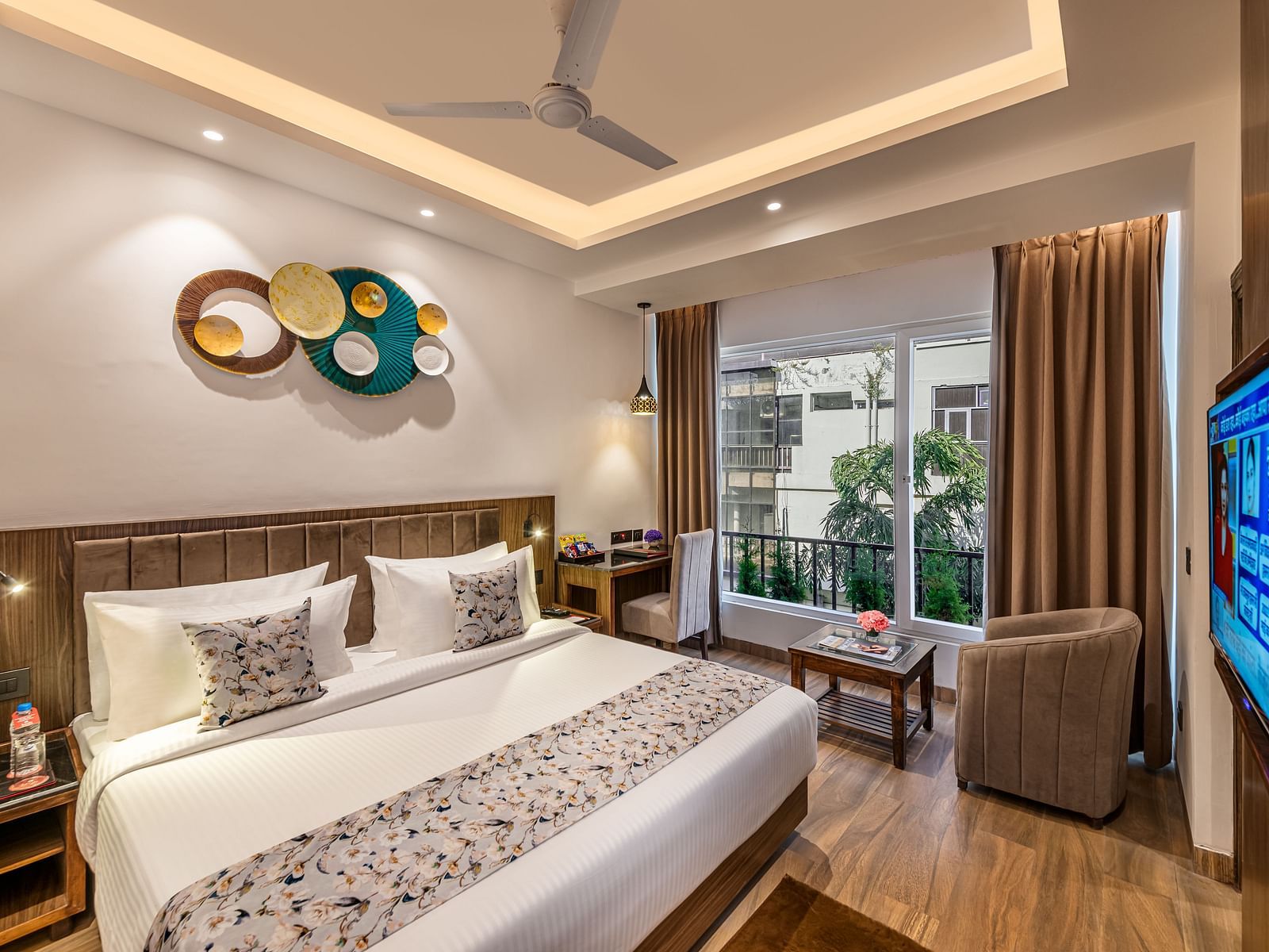 A bright, modern hotel room with a king size bed, decorative wall art, a brown armchair, small table, flat-screen TV, and an open door leading to a balcony - Lords Eco Inn Dehradun.