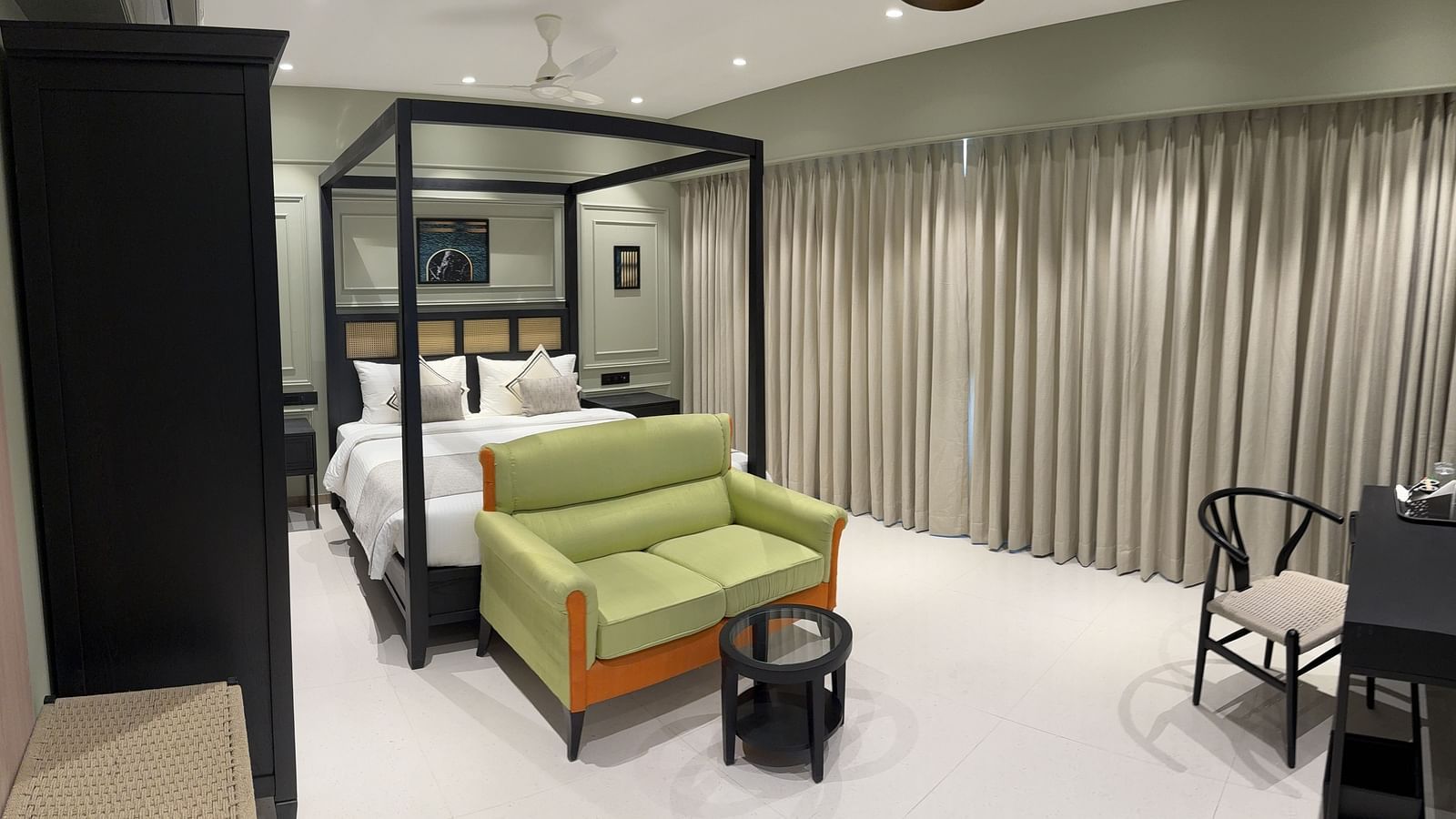 A spacious hotel room with a black four-poster bed, white bedding, a lime green sofa, a black round coffee table, and two black armchairs, all under a white ceiling fan and recessed lighting.