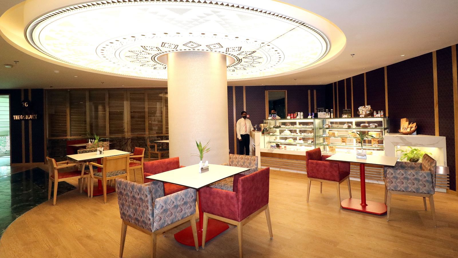  Brown and cream Swosti Premium with an oval ceiling light, beneath which tables and chairs are arranged, featuring neatly set tables.