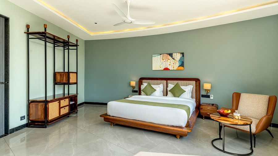 Pool View Room featuring a king size bed with a green and white patterned bedspread, wooden furniture, and a large window with sheer curtains at The Qualia Resort Club & Brewery, Udaipur.