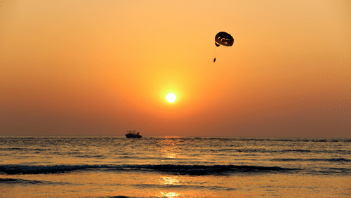 An image of beach where a parachute can be seen attached to a boat with the backdrop of sunset