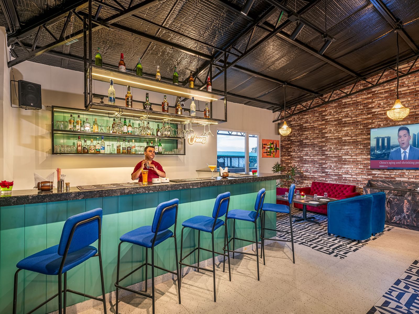 A bar with a bartender behind the counter, blue bar stools, and a selection of bottles on display. It features a high ceiling with exposed beams, a brick wall, and a lounge area with blue seating - Lords Eco Inn Dehradun.